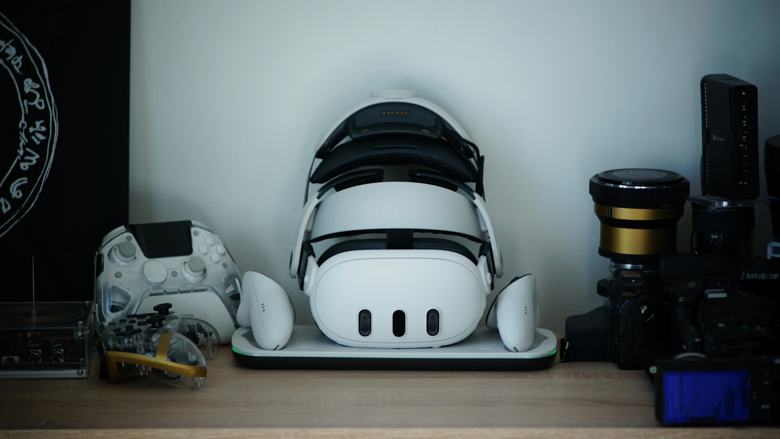 White standalone VR headset and controllers on a charging dock on a wooden shelf beside game controllers and cameras